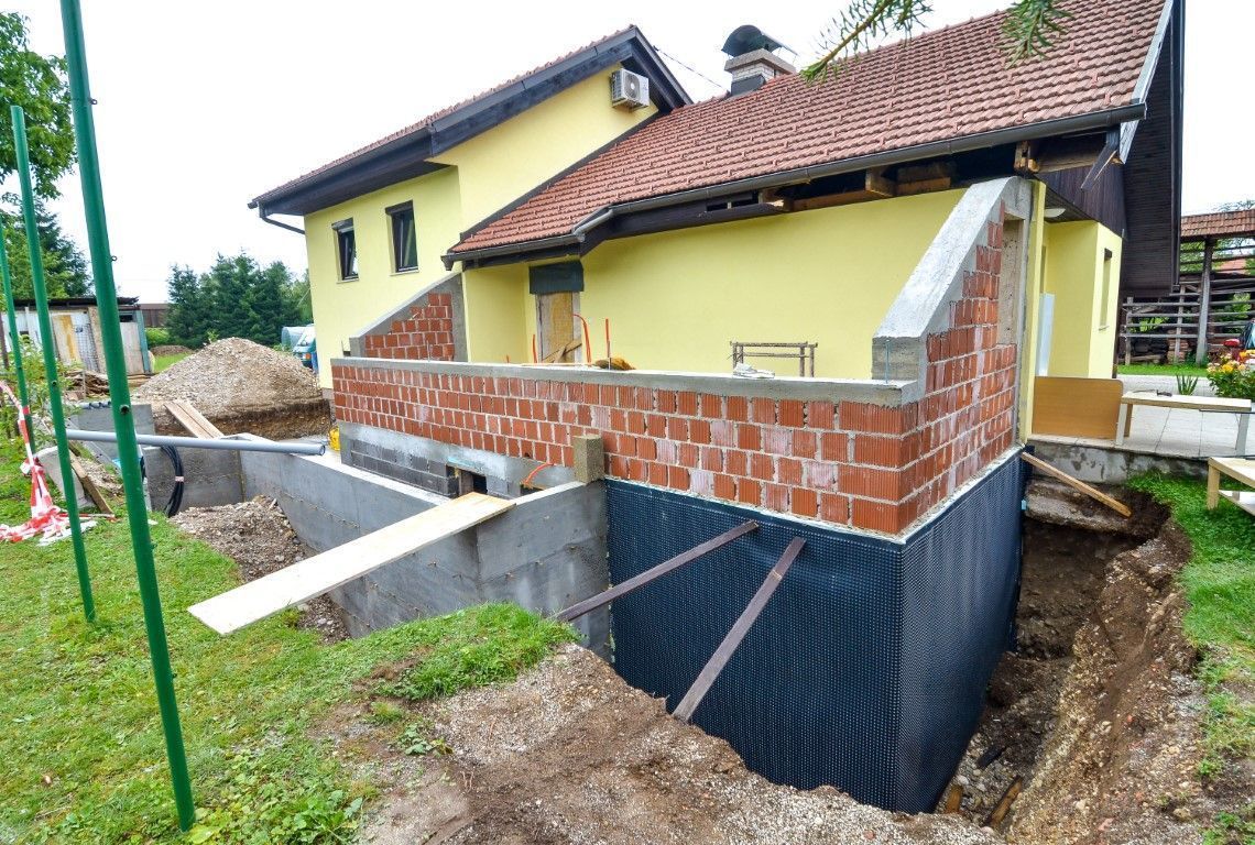 Partially renovated house with red roof, yellow walls, and brick foundation work in a muddy yard