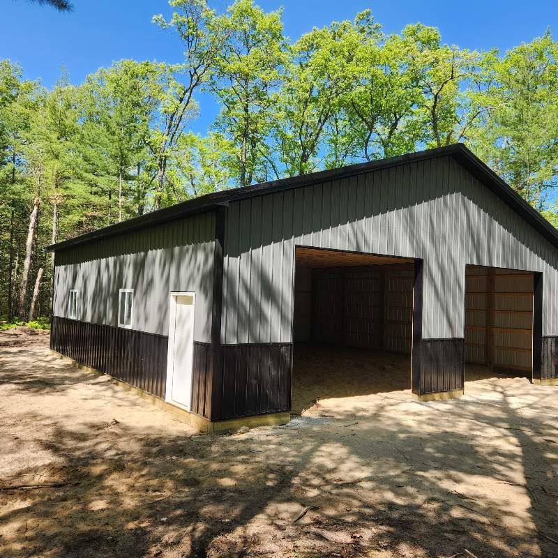 A newly constructed metal pole barn with grey upper walls and black lower siding, set in a wooded area under a blue sky.