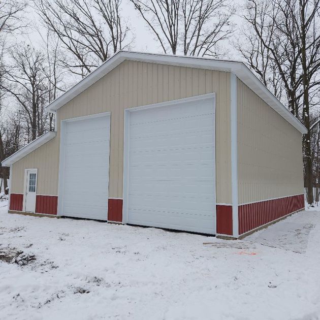 A tan and red metal pole barn with two white garage doors and a side entry door, set in a snowy, wooded environment.