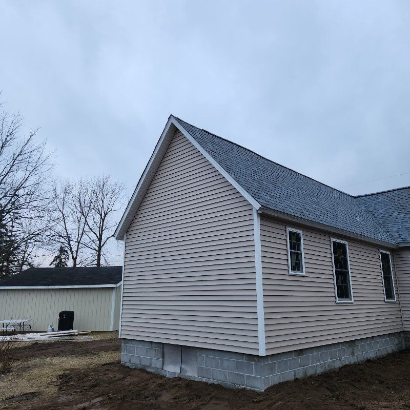 A newly constructed house with beige vinyl siding and a gray shingled roof, set on a block foundation against a gray sky.