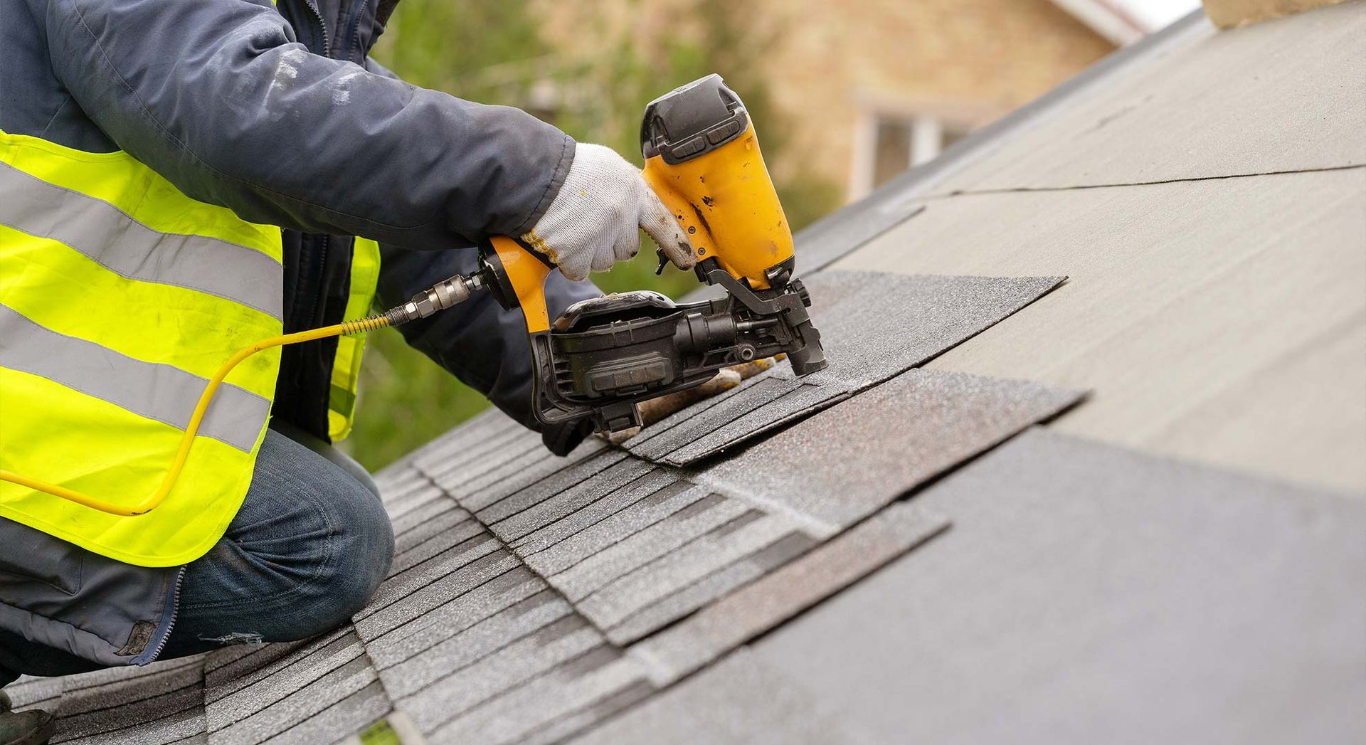 A worker in a high-visibility vest uses a yellow pneumatic nail gun to install shingles on a residential roof.