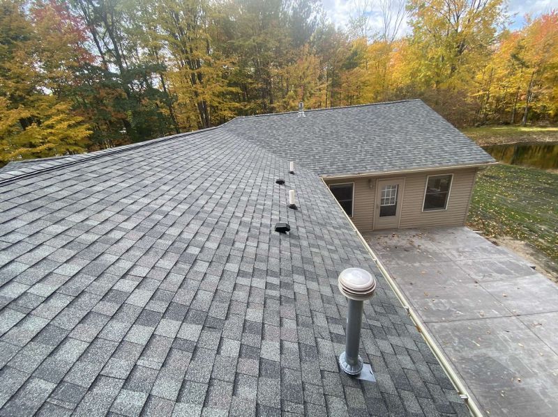 High-angle view of a grey shingled roof with several vents, overlooking a concrete patio and a wooded area in autumn.