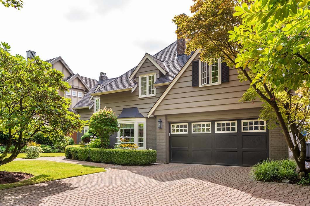 A suburban house with a brown exterior, a gray shingled roof, a dark two-car garage, and a patterned brick driveway.
