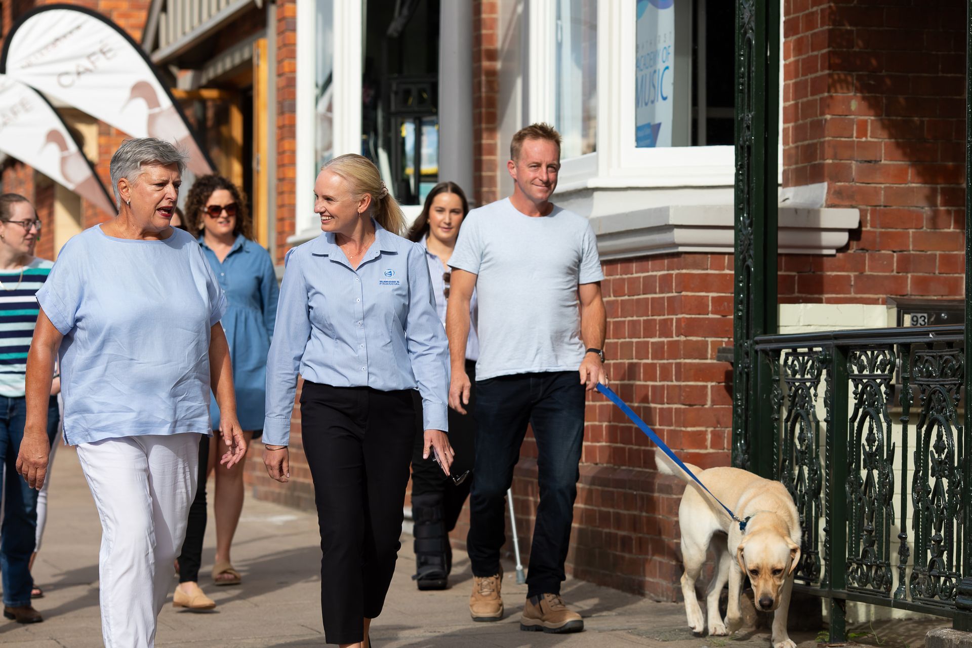 Four people walk along a street; a man in front leads a guide dog on a leash.