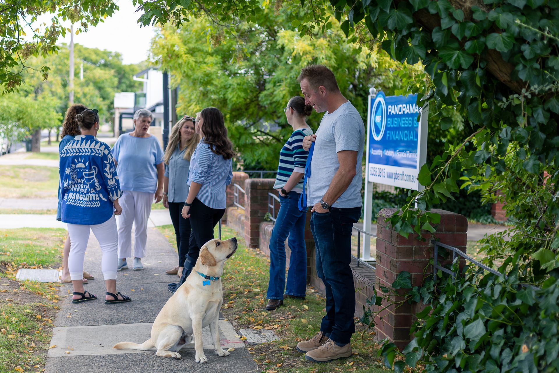 People talking on a sidewalk by a sign, with a seated dog in the foreground.