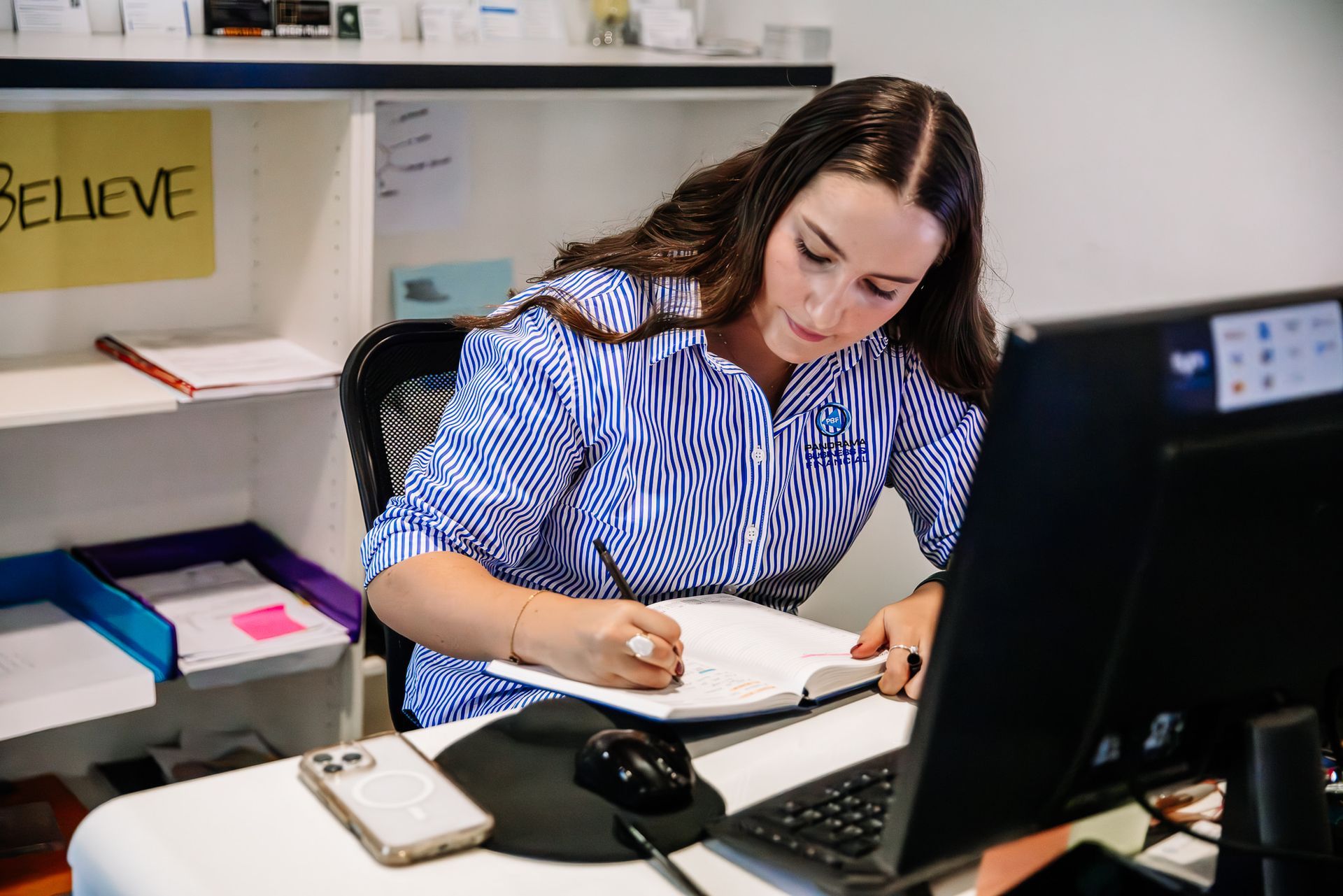 Woman writing in a notebook at a desk with a computer monitor in an office