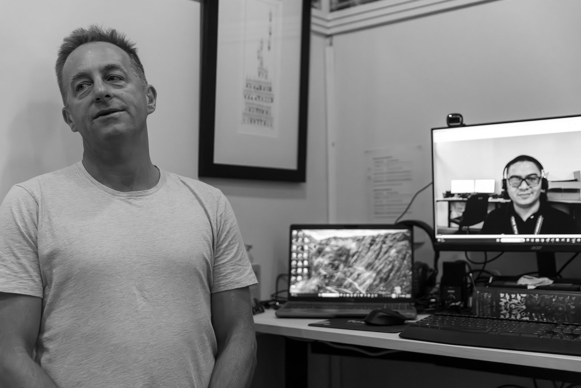 Man standing beside desk with laptop and monitor in a small office, looking slightly upward