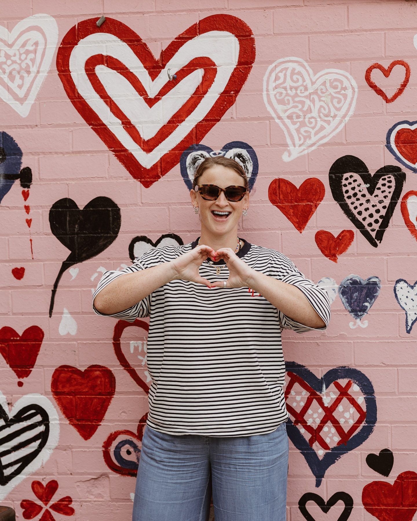 Person in sunglasses making a heart shape with hands in front of a pink heart-painted wall