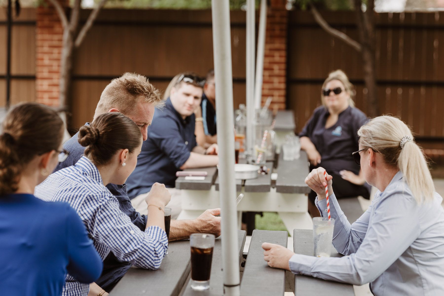 Group of coworkers in an outdoor meeting, seated around a long picnic table and talking over drinks.