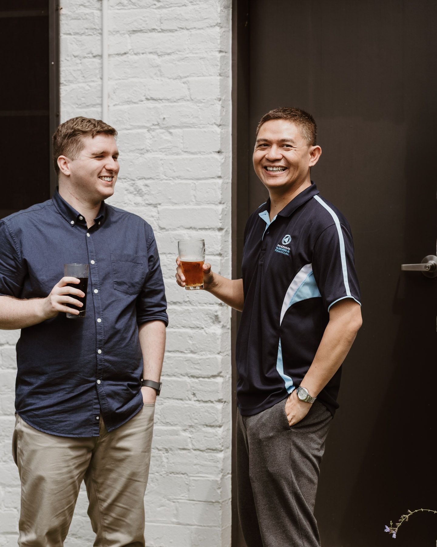 Two people standing by a white brick wall, smiling and holding drinks outside a building.