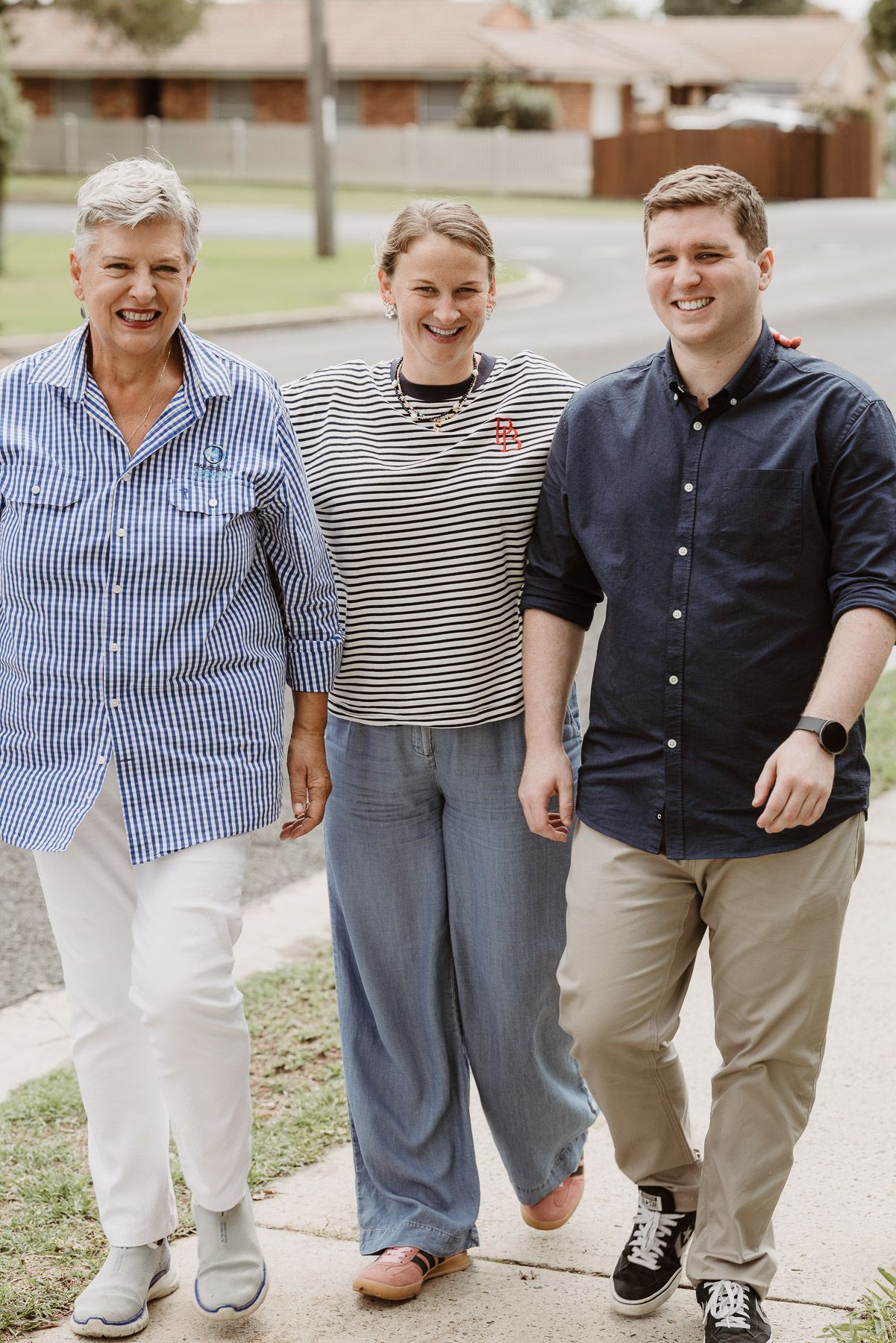 Three people walking together on a sidewalk in front of suburban houses, smiling.