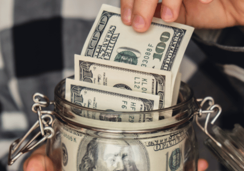 Hand placing US dollar bills into a glass jar, representing saving money and budgeting.