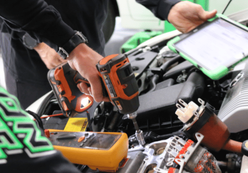 Technician using power tool and tablet to work on a car engine, performing diagnostics and repairs.