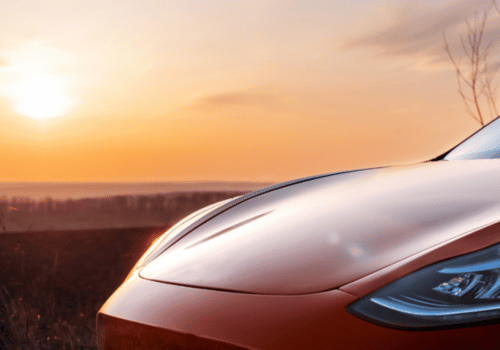 Close-up of a red Tesla hood and headlight at sunset, with warm light reflecting over a calm view.