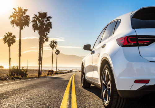 White SUV driving along a coastal road lined with palm trees at sunset overlooking the ocean.