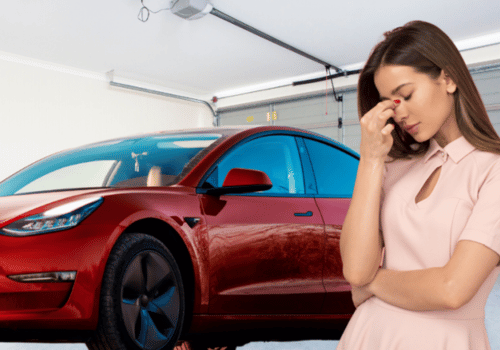 Woman looking stressed in a garage beside a red car.