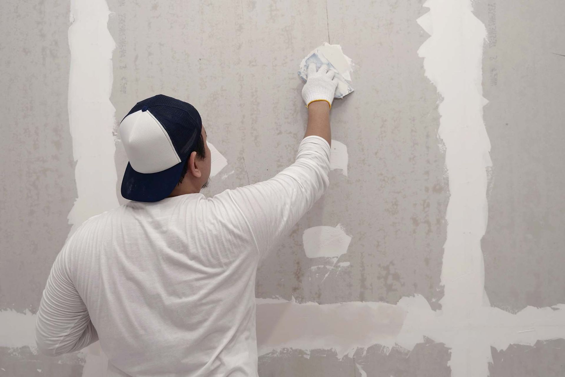 Person in white shirt and cap applies plaster to a wall with a trowel.