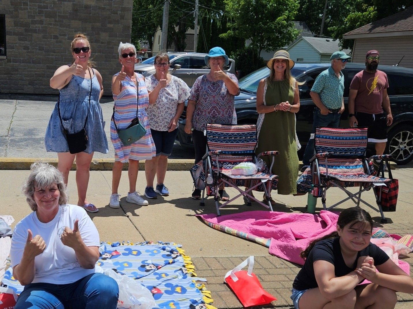 A group of people are sitting on blankets and chairs on the sidewalk.