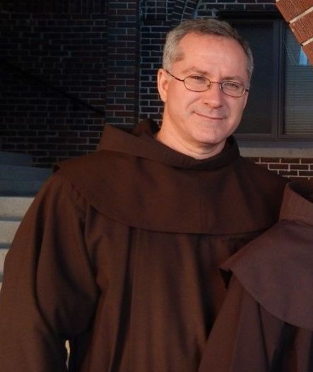 A man wearing glasses and a brown robe stands in front of a brick building
