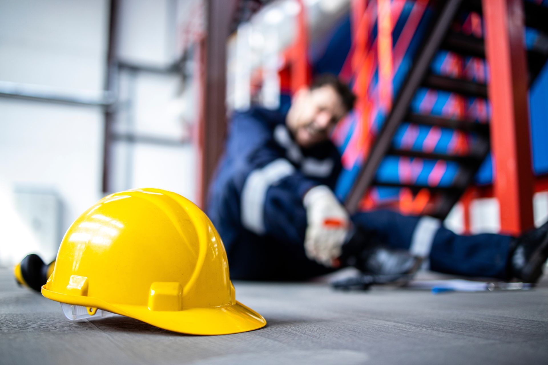 Yellow hard hat on floor; worker in blue uniform, fallen near a ladder in a factory.