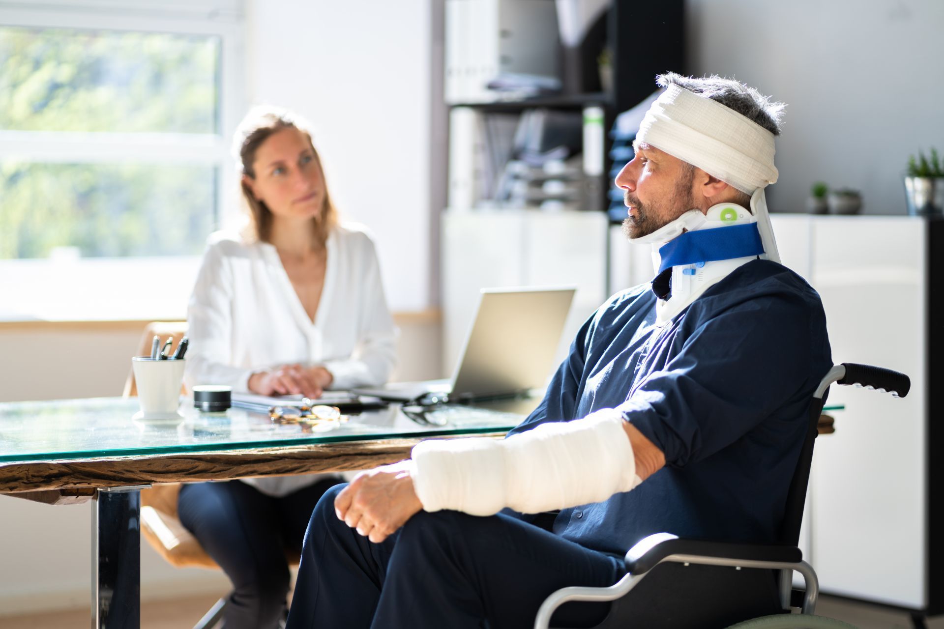 Man in wheelchair with bandages, neck brace, and cast, talking to a woman at a desk in an office.