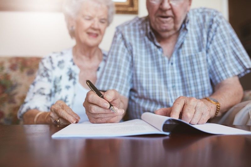 Senior couple signing documents at a table; woman smiling, man looking on.