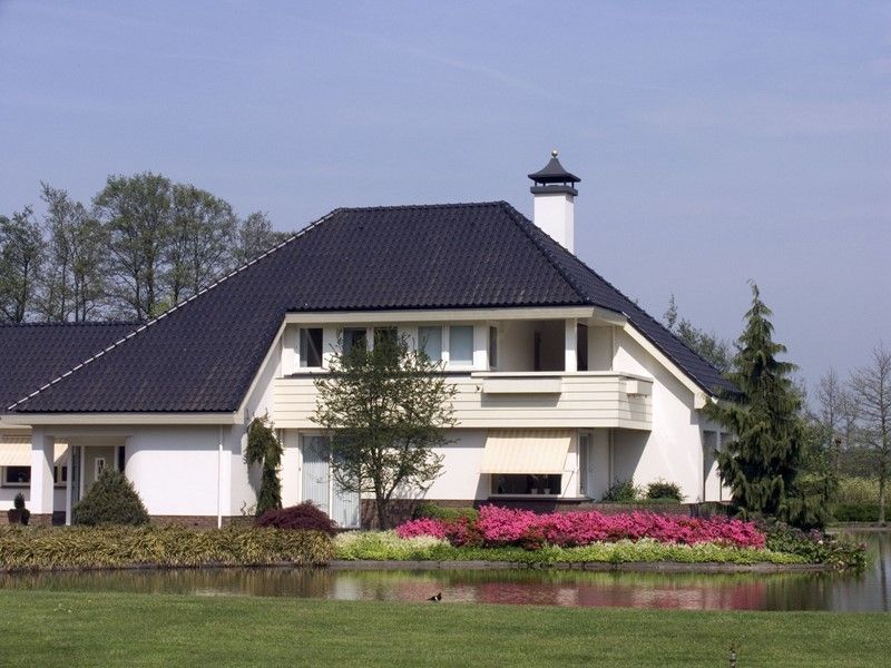 White house with black roof, balcony, and chimney, next to a pond and flower bed under a blue sky.