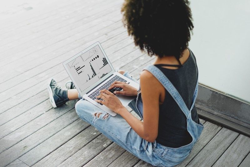 Person seated on wooden surface, using laptop. Wearing overalls and a dark shirt.