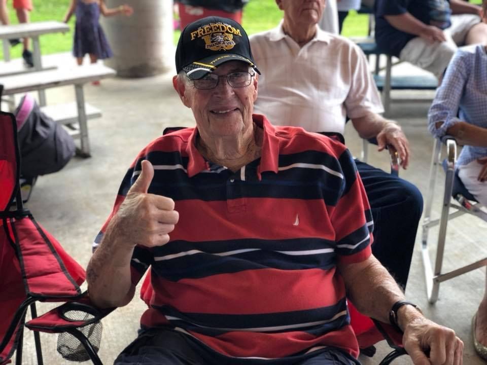 An elderly man is giving a thumbs up while sitting in a chair.