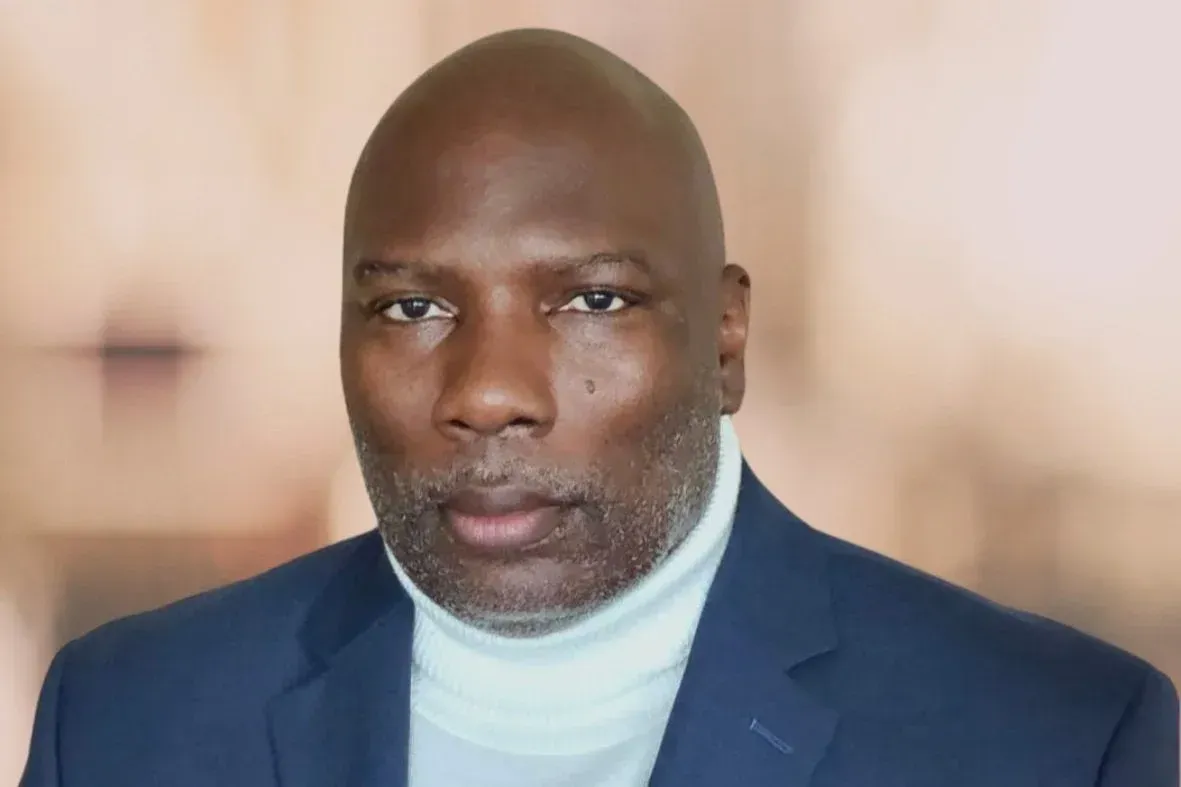 A person wearing a blue blazer and a light-colored turtleneck sweater, posed against a blurred, neutral background.