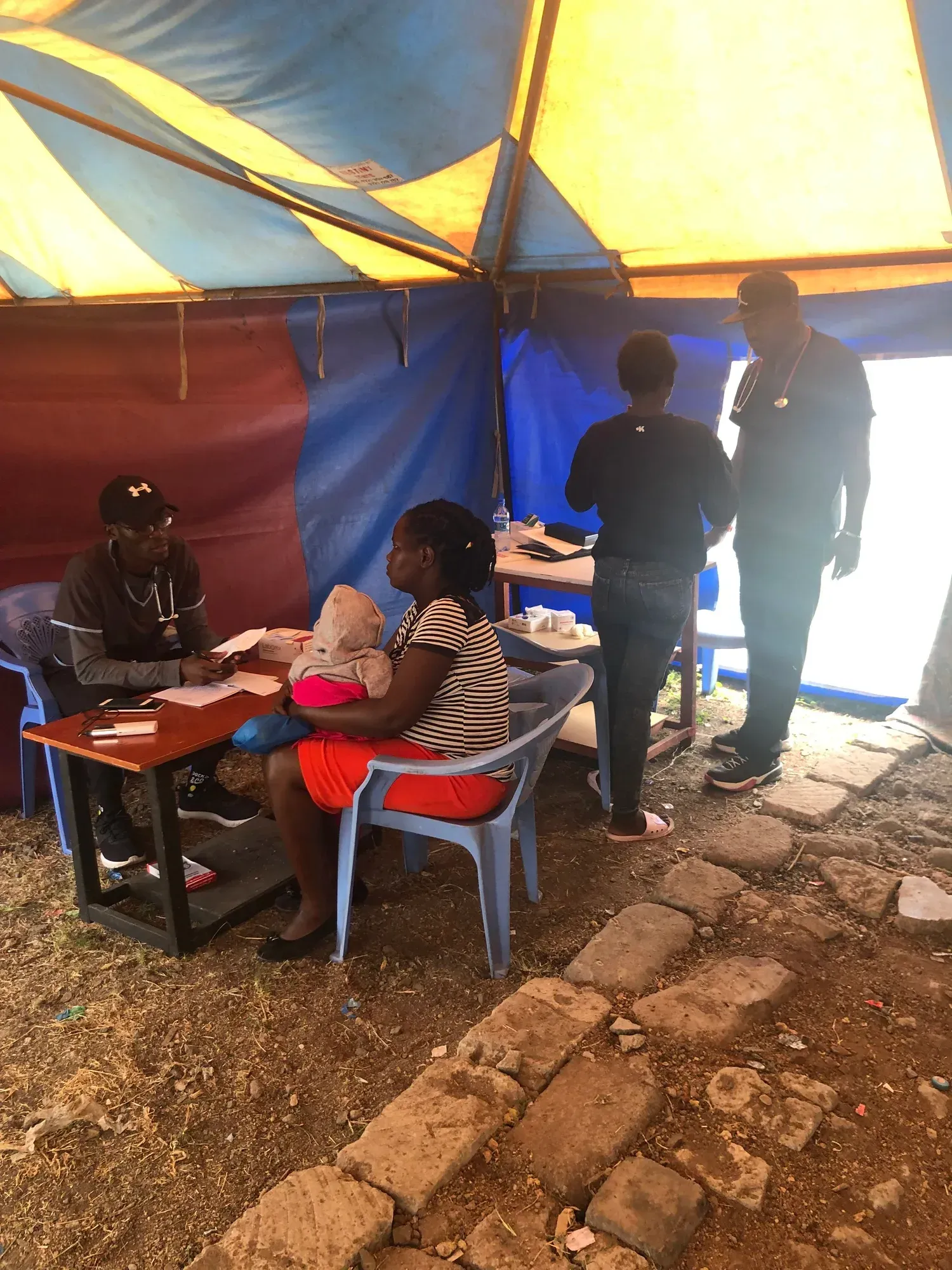 An indoor clinic scene inside a tent with a health worker consulting a patient holding a baby while others stand nearby.