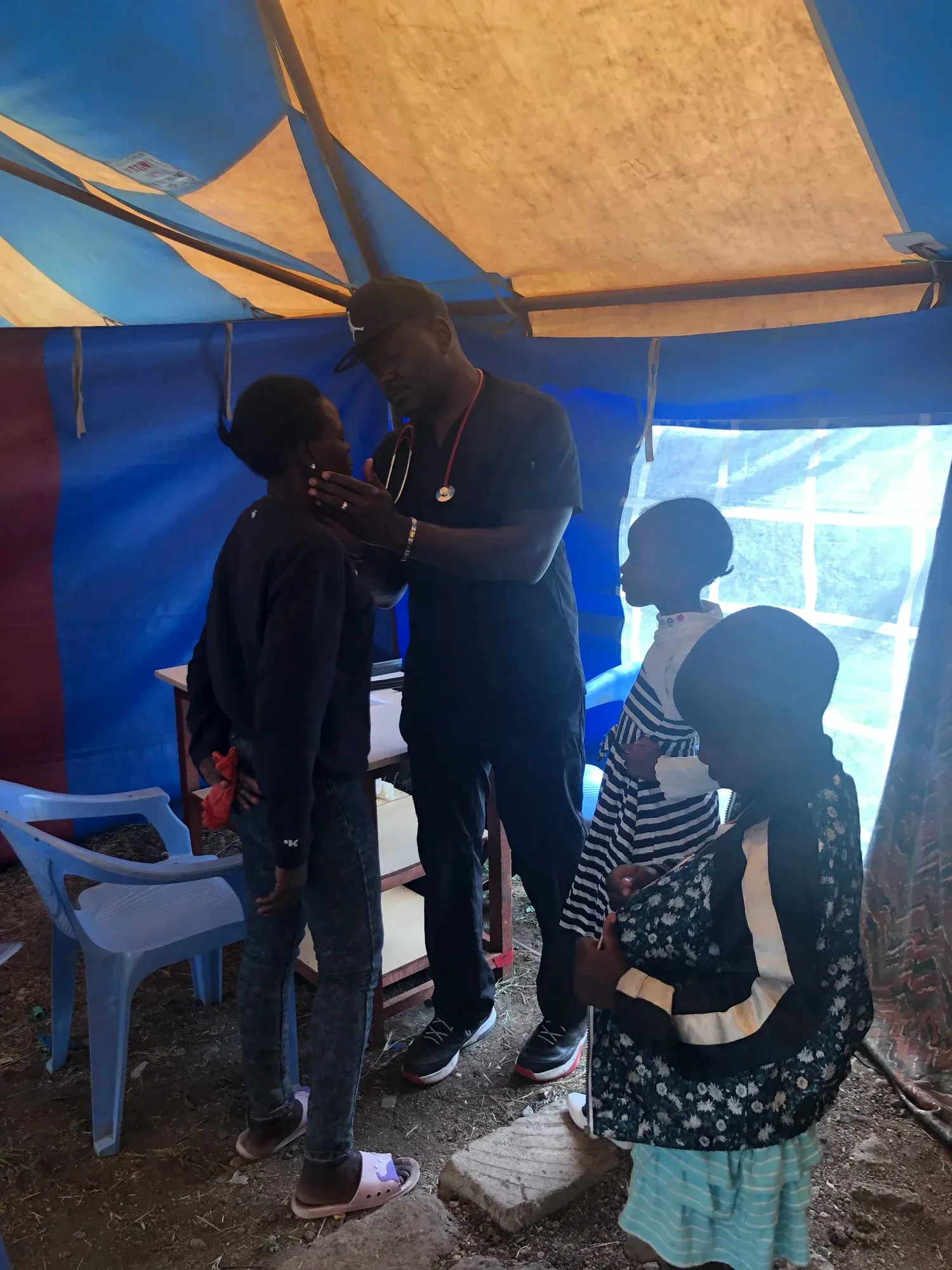 A healthcare provider in a tent checks a patient’s face, while two individuals stand by waiting.