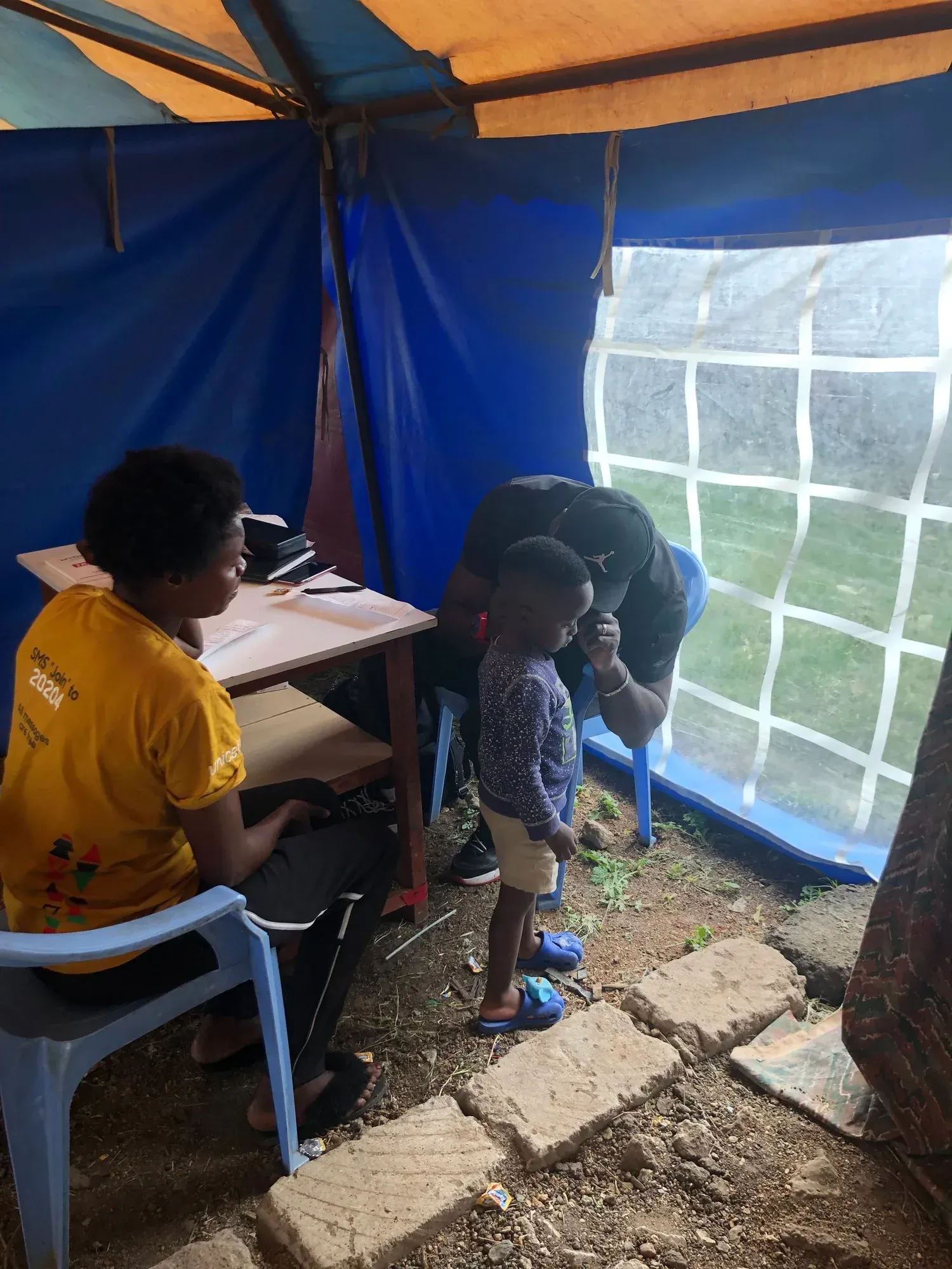 A child stands in a tent while being examined by a person wearing a black cap; a person sits at a desk nearby.