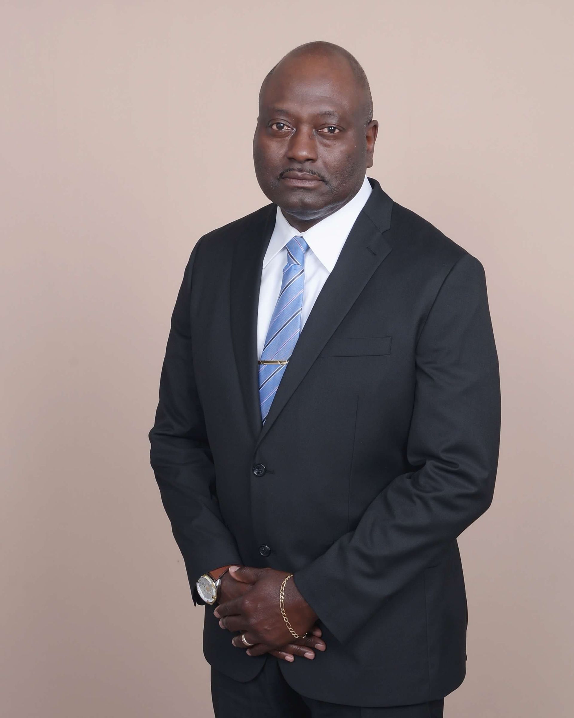 Headshot of a person with a short beard, wearing a dark patterned blazer and white shirt against a neutral background.