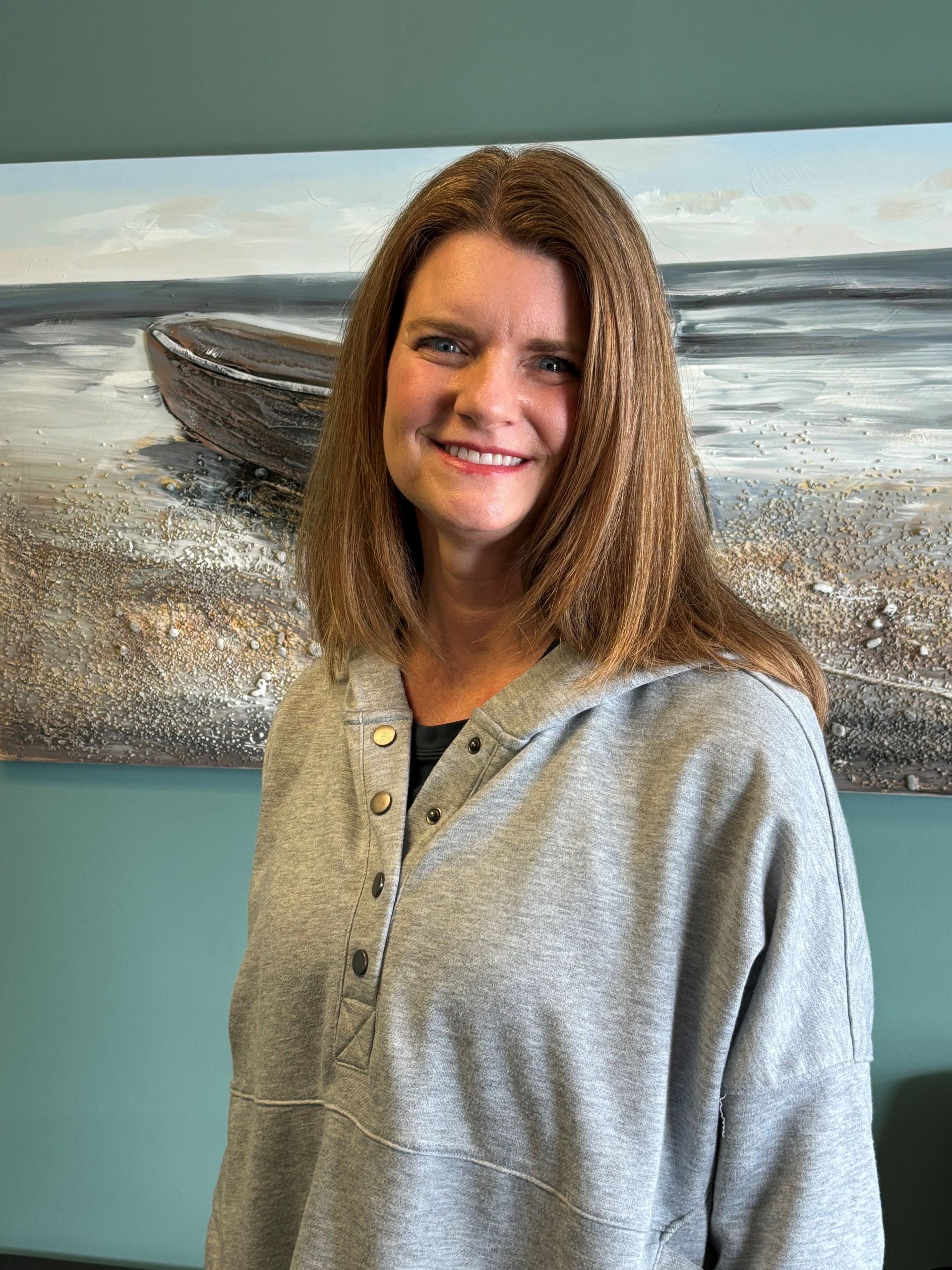 A woman is smiling in front of a painting of a boat on the beach.