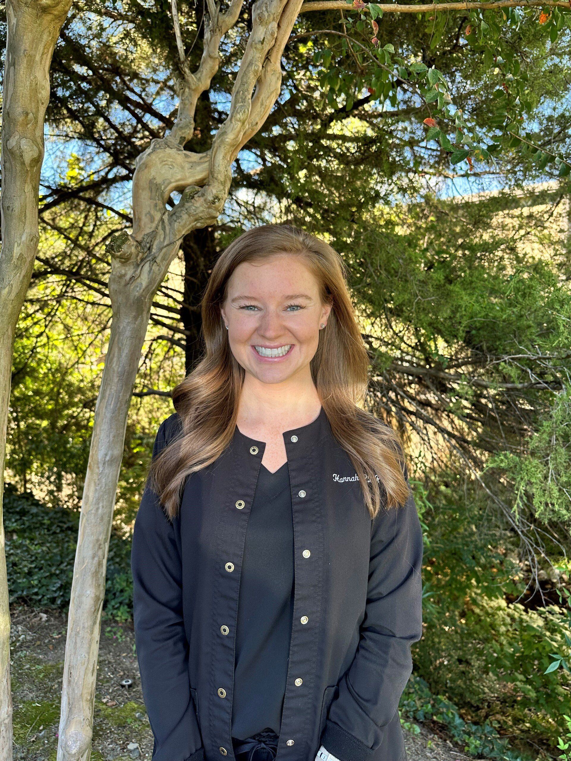A woman in a black jacket is smiling in front of a tree.