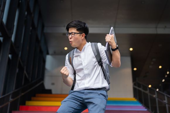 A man with a backpack is standing on a set of rainbow colored stairs. A man with a backpack is standing on a set of rainbow colored stairs.