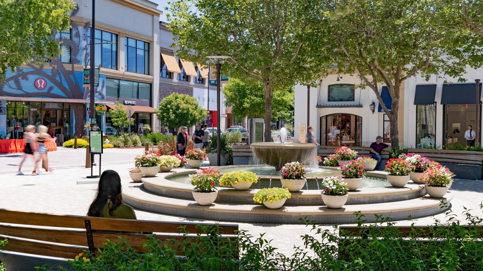 Fountain in a shopping area, surrounded by flowerpots. People walk and sit nearby in the sunshine  at The Waymark in Walnut Creek, CA.