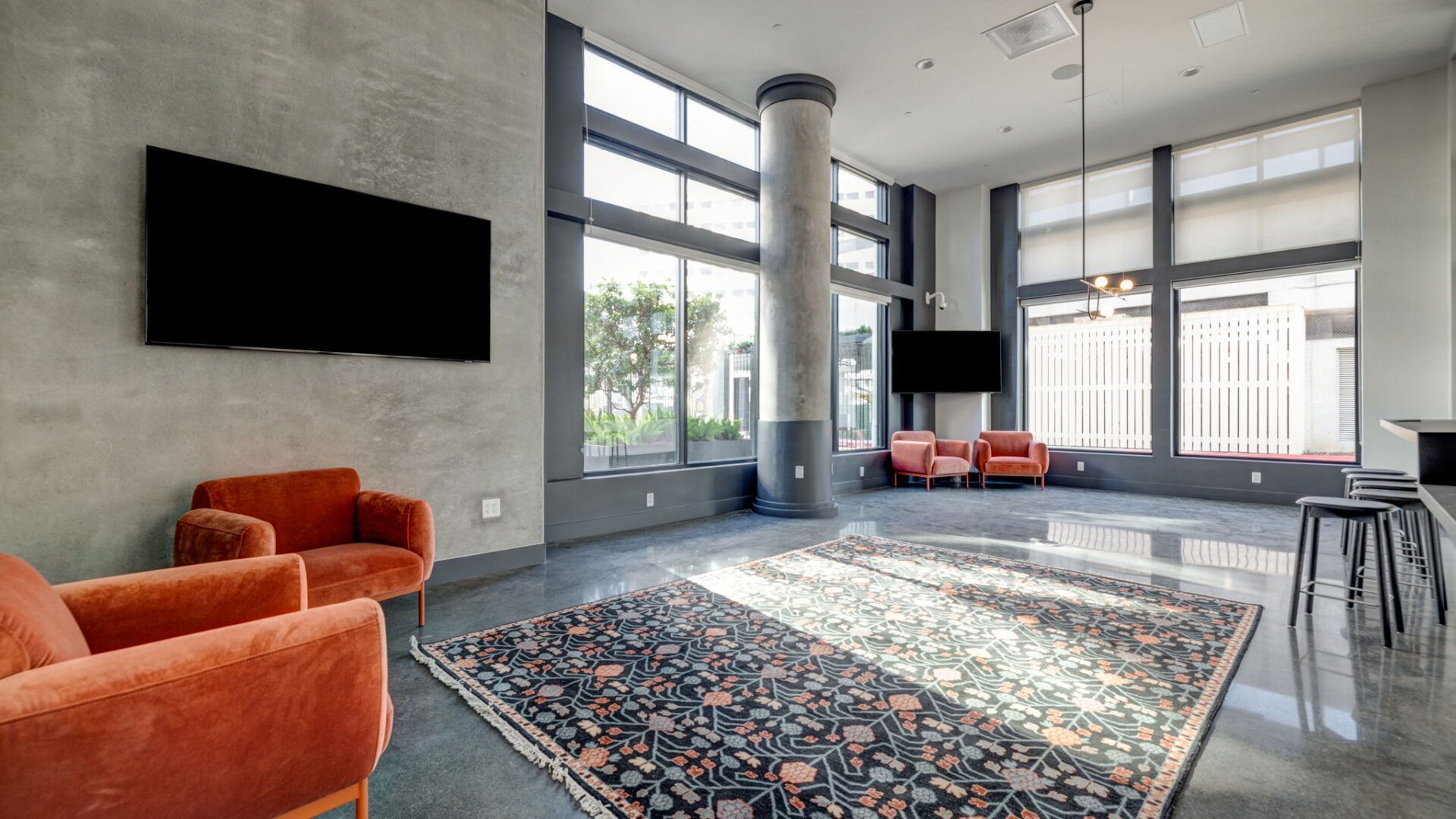 Lounge with orange chairs, TVs, and rug, near windows and a concrete pillar in a modern building at The Waymark in Walnut Creek, CA.