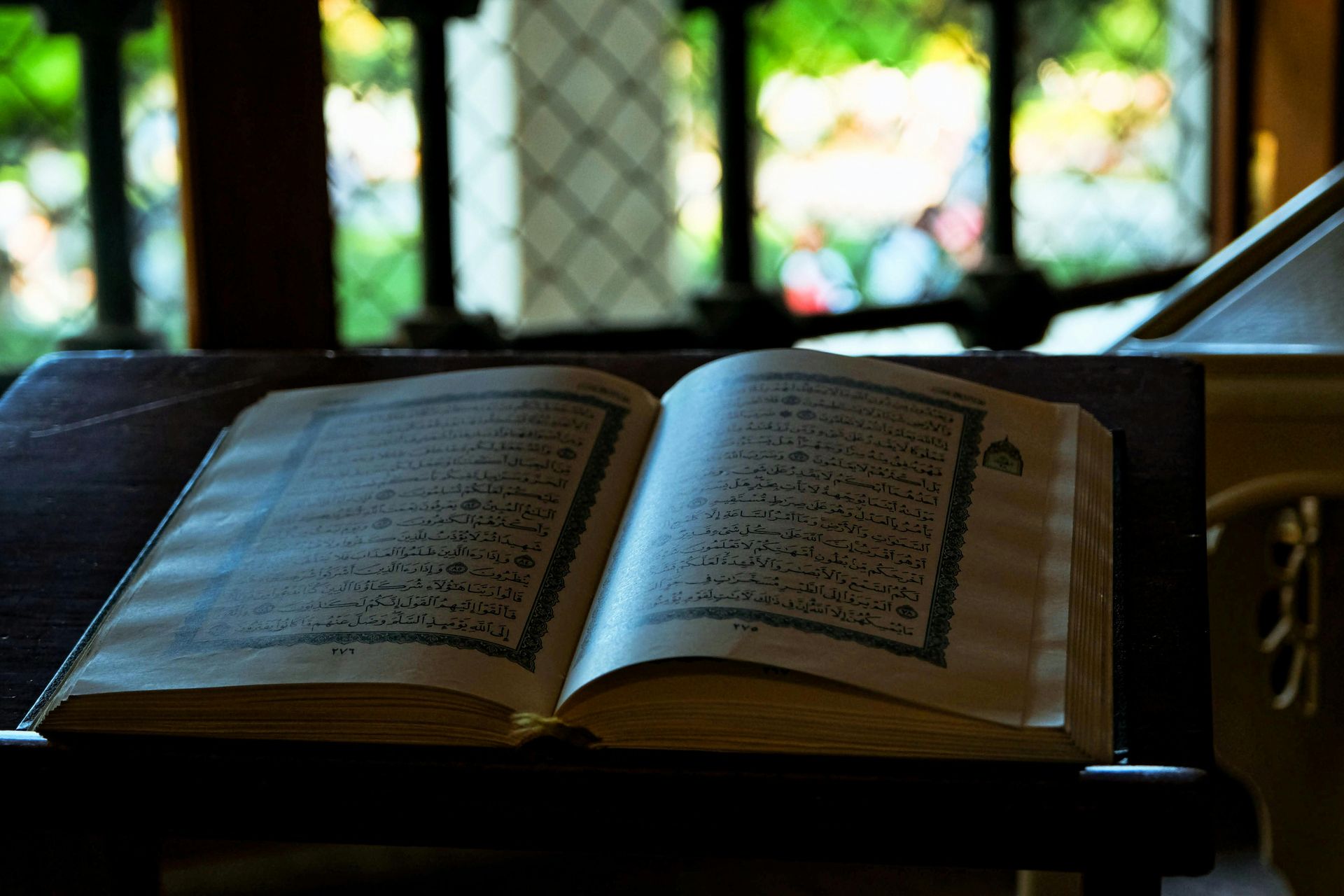 An open Quran rests on a wooden stand in front of a window with metal bars.