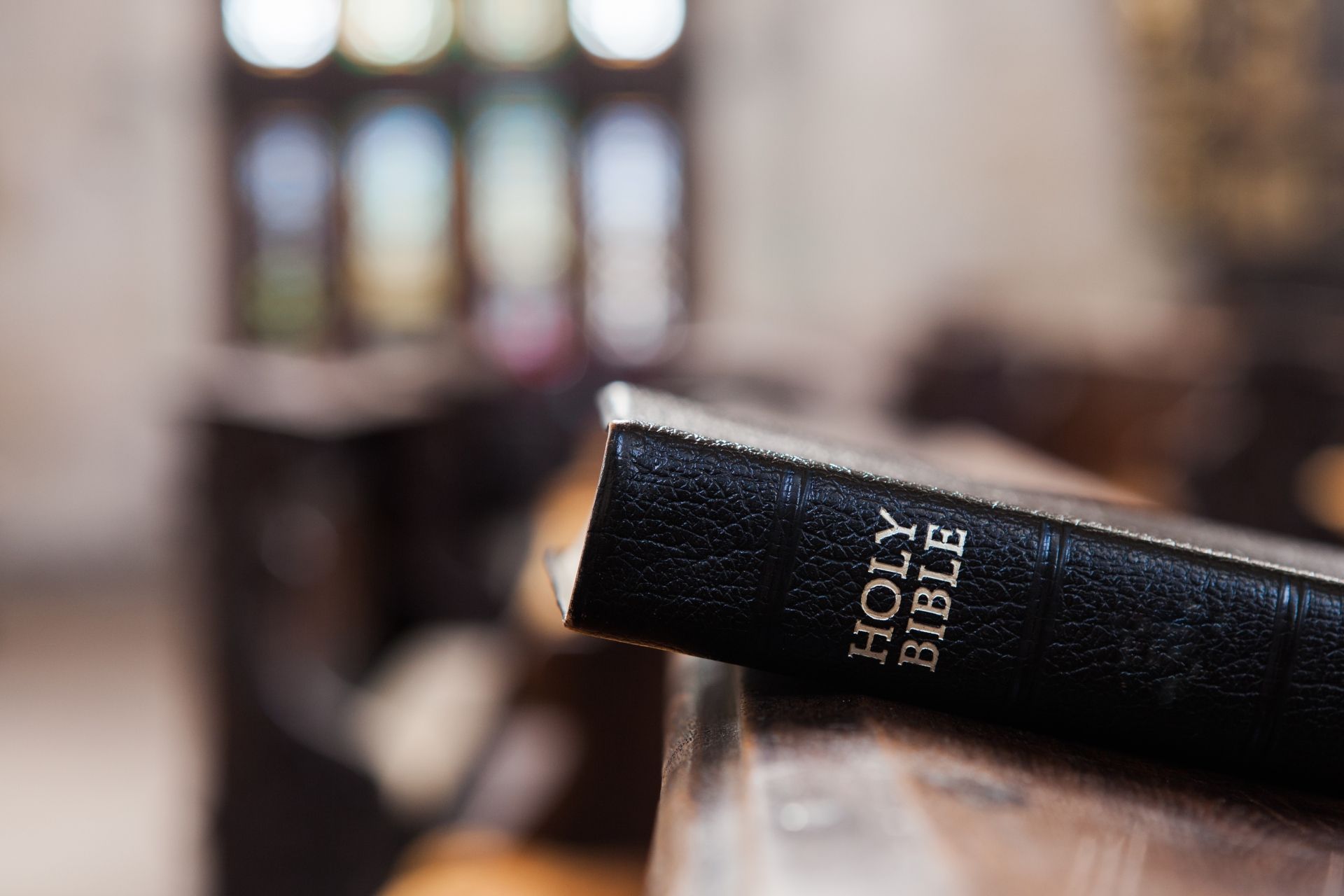 A black Holy Bible sits on a wooden pew inside a sunlit church.