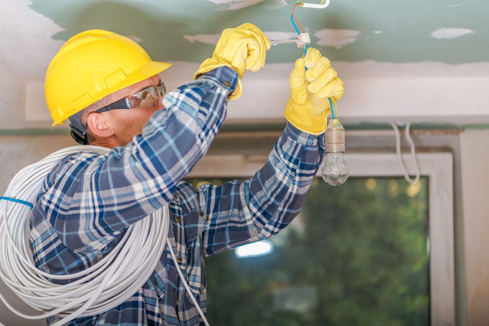 An electrician wearing a hard hat, safety glasses, and gloves, installs a light fixture from a ceiling in an interior room.