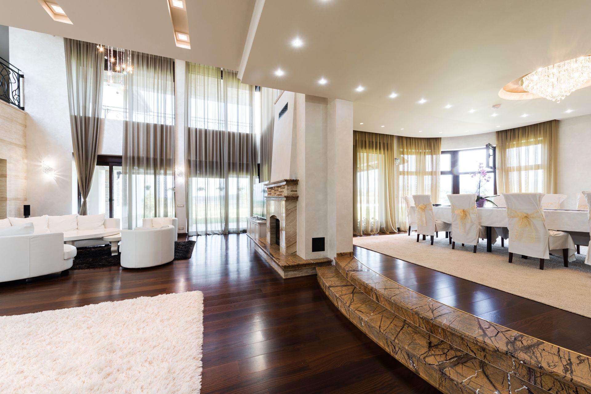 A spacious, high-ceilinged living and dining area with hardwood floors, white furniture, and large windows with curtains.