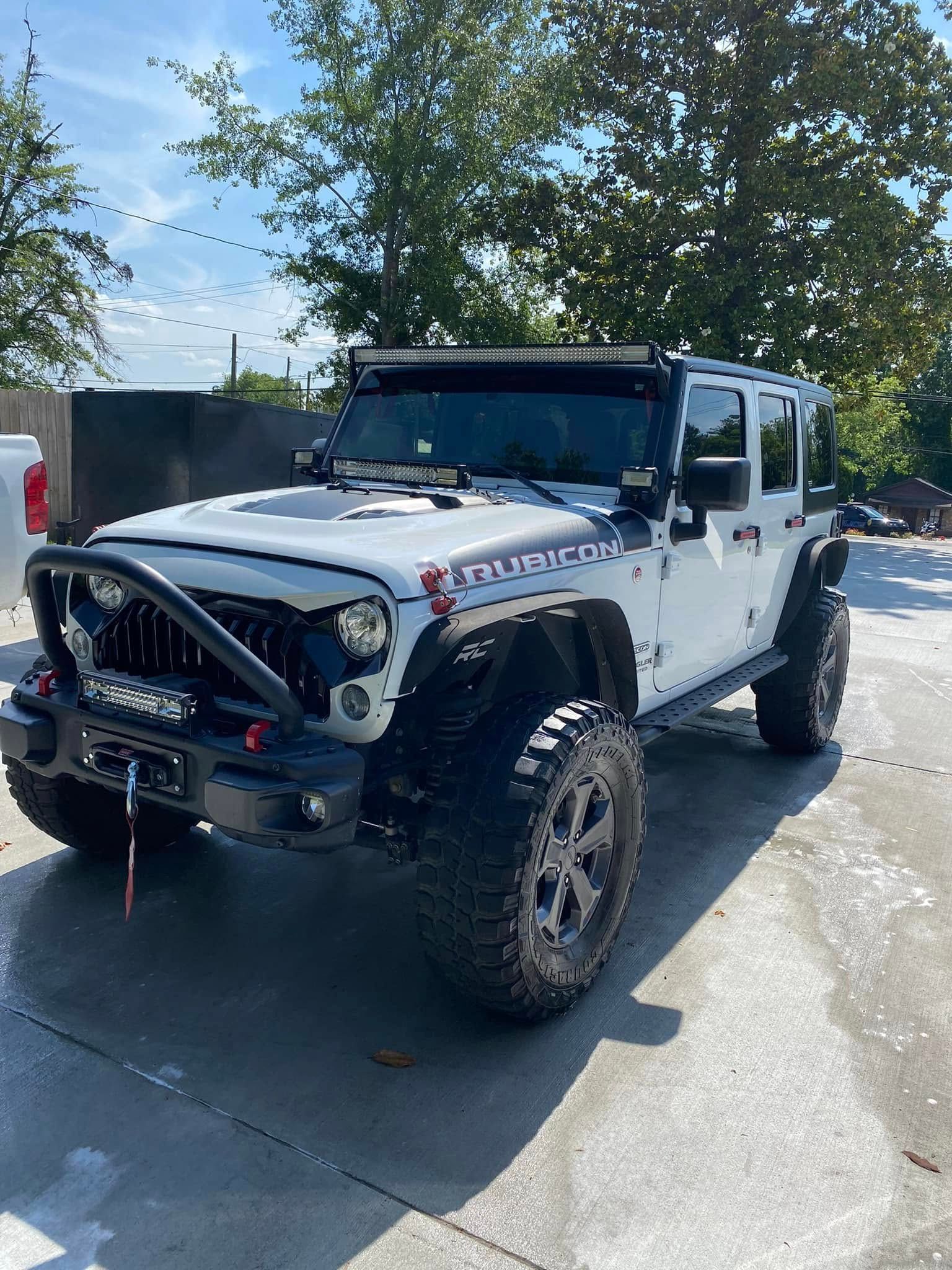 White off-road Jeep Wrangler with lifted tires and front grille guard, parked outdoors on a sunny day