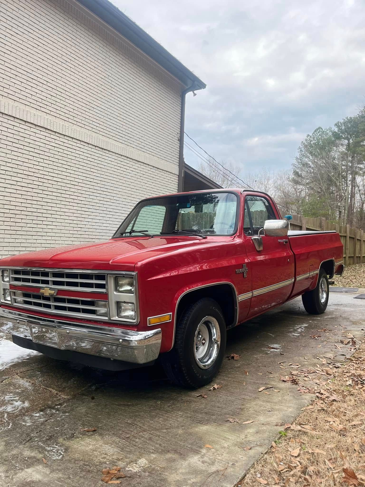 Red vintage pickup truck parked beside a white building on a wet driveway.