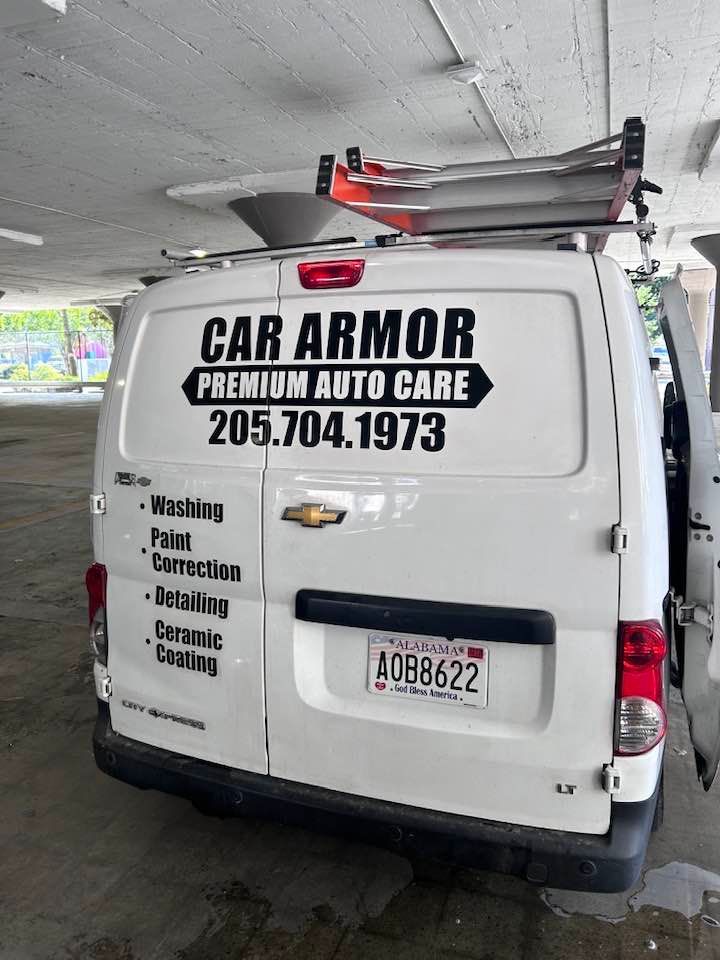 White CAR ARMOR auto care van in a parking garage, rear view with roof ladder and large business logo.
