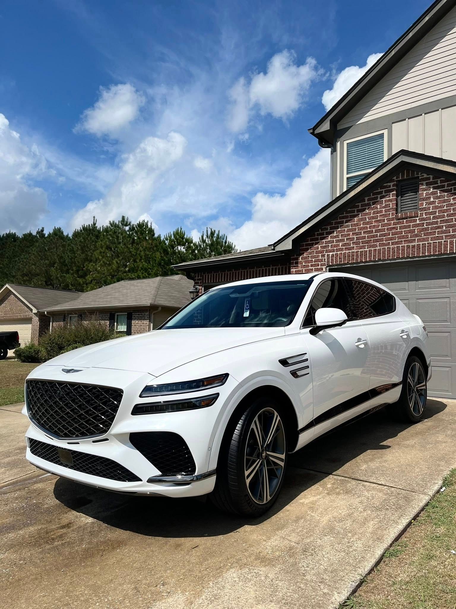 White SUV parked in a driveway beside a brick house under a blue sky with clouds