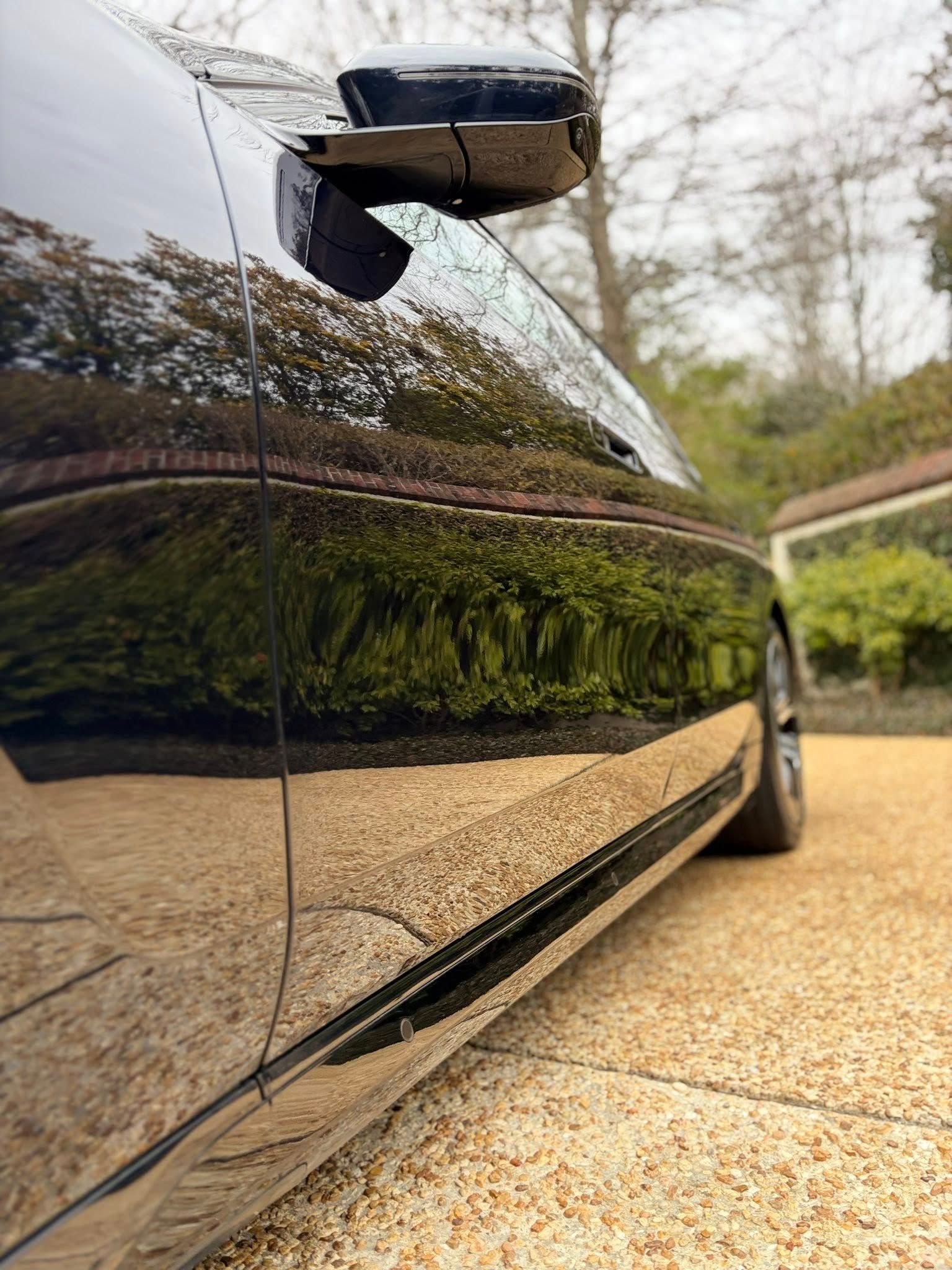 Close-up of a black car’s glossy side panel and mirror reflecting trees and a gravel driveway
