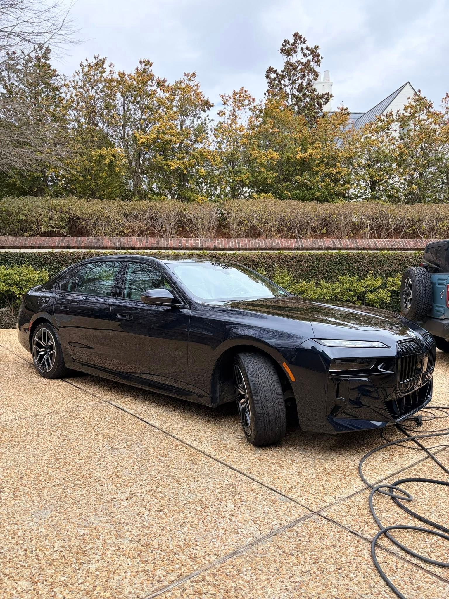 Black sports sedan parked on gravel, viewed from the front-left with trees and a hedge behind it.