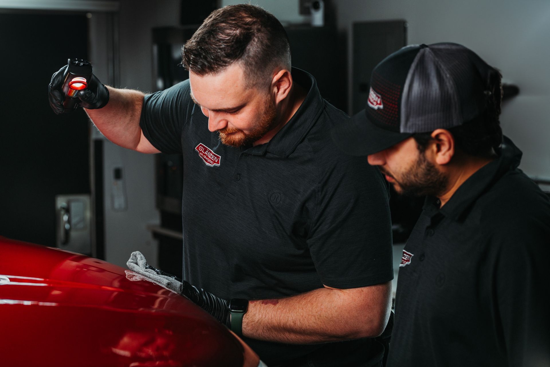 Two men are working on a red car in a garage.